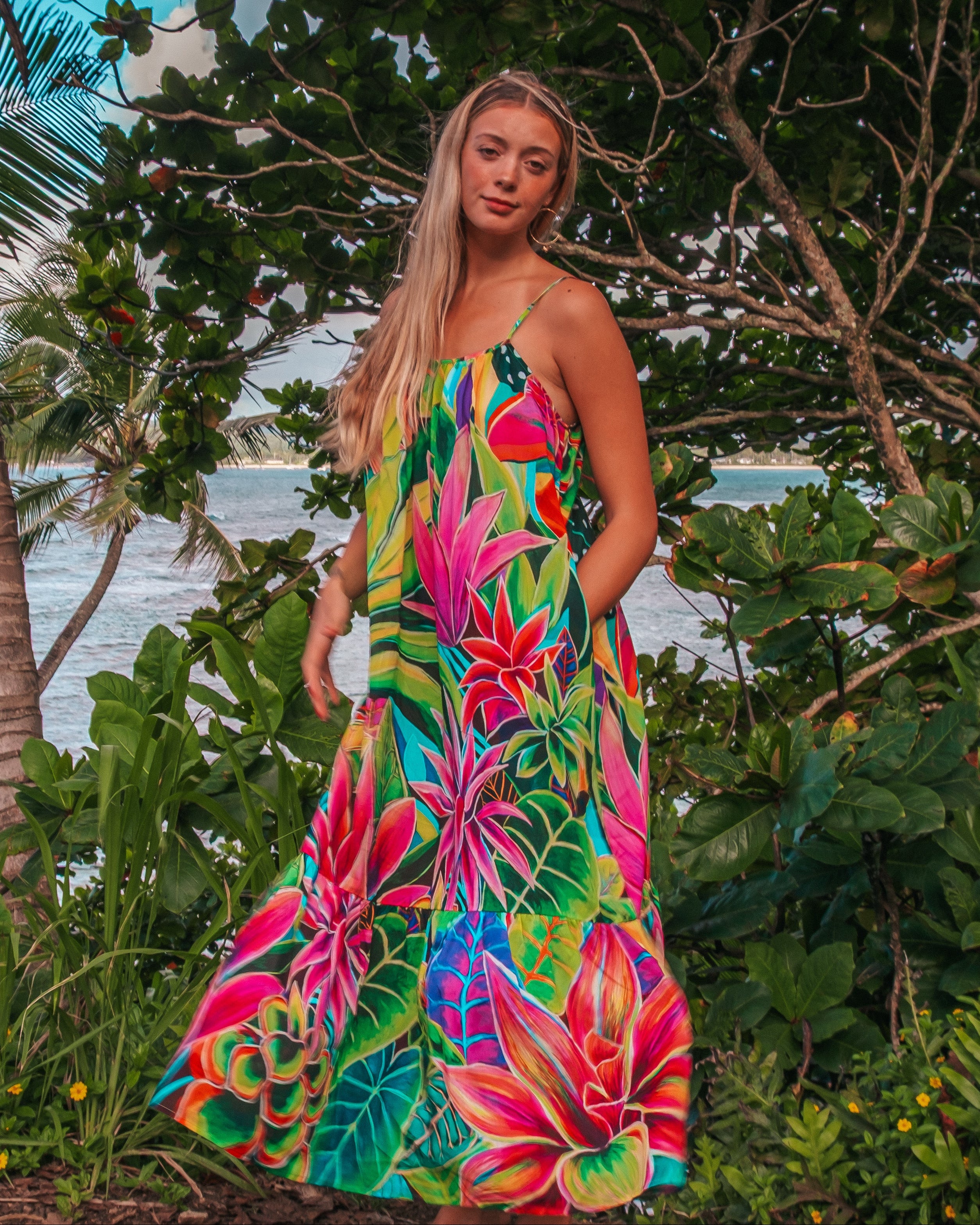 A woman stands outdoors in a tropical garden with palm trees and ocean scenery in the background, wearing a vibrant strapless maxi dress with a colorful floral and leaf print.