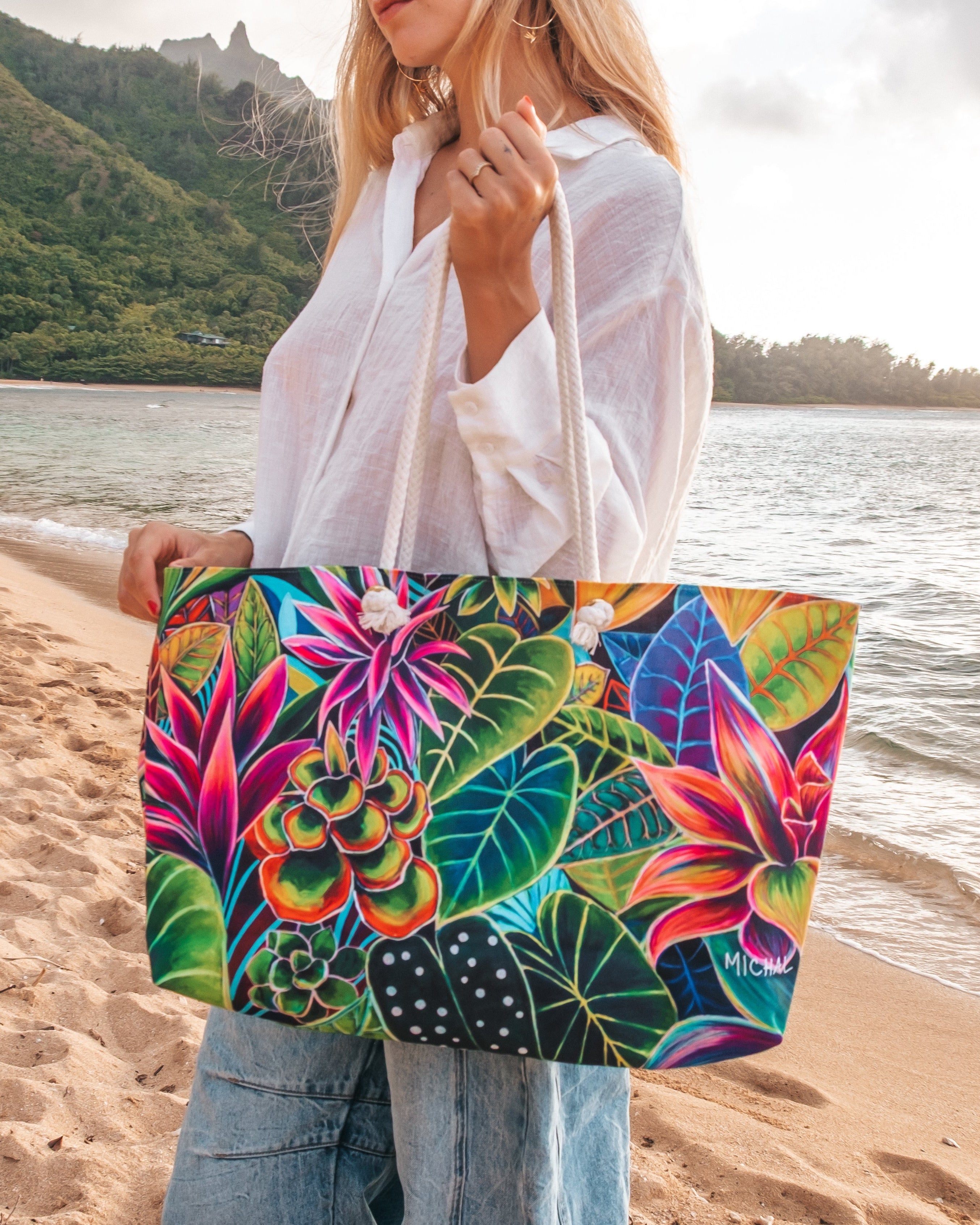 A woman standing on a sandy beach holds a large tote bag with a vibrant, multicolored tropical floral and leaf print. The background features the ocean, distant green hills, and a cloudy sky.