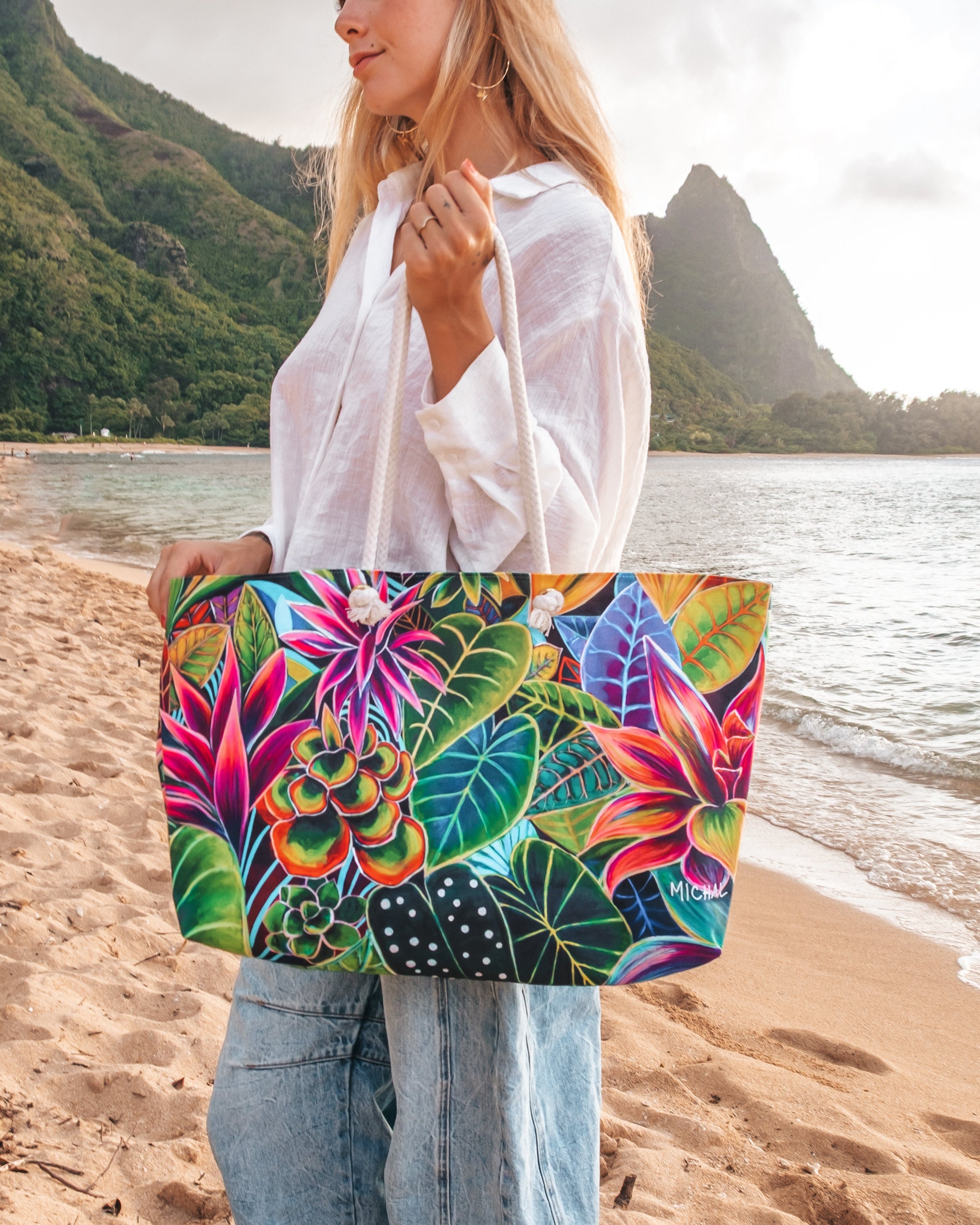 A woman standing on a sandy beach holds a large tote bag with a vibrant tropical floral and leaf print. The background features a scenic ocean view with green mountains and a cloudy sky.